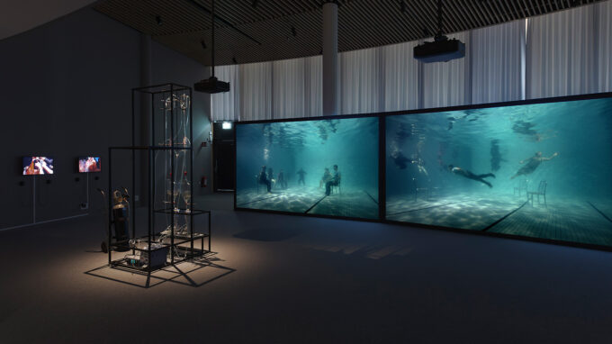 Yta med tvåstora bildskärmar som visar människor under vattnet. Konstverk från Borderlands.